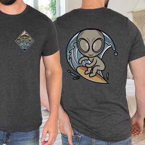 May include: A dark gray t-shirt with a graphic of a gray alien riding a surfboard on a wave. The alien has large eyes and a small mouth. The wave is blue and white. The t-shirt has a small graphic of a UFO on the left chest.