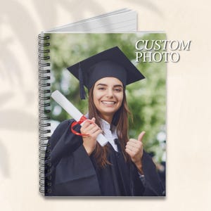 May include: A black graduation cap and gown with a red ribbon. The person is holding a diploma and giving a thumbs up. The background is a blurred green.