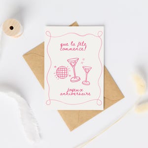 Könnte beinhalten: Eine weiße Grußkarte mit rosa Illustrationen einer Discokugel und zwei Martinigläsern, mit den rosa Schriftzügen "que la fête commence!" und "joyeux anniversaire". Die Karte liegt auf einem braunen Umschlag.