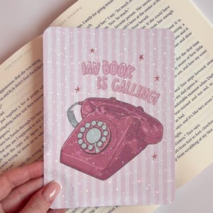 Op de afbeelding: Roze en wit gestreept notitieboek met de tekst "MY BOOK IS CALLING!" en een roze draaischijftelefoon illustratie. Het notitieboek wordt opengehouden, met een boek op de achtergrond.