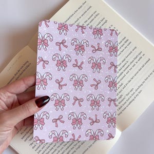 May include: A rectangular card featuring a repeating pattern of pink candy canes and bows on a light pink background. The candy canes have white and pink stripes, and the bows are pink. The card is held over an open book.