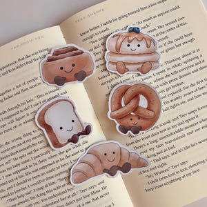 Op de afbeelding: Een verzameling van vijf stickers met een voedselthema, waaronder een kaneelbroodje, pannenkoeken met bosbessen, een krakeling, een sneetje toast en een croissant. Elke sticker heeft een lachend gezicht en kleine armen en benen.