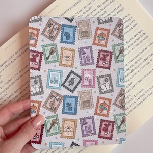 Könnte beinhalten: Ein quadratischer Lesezeichen mit einem sich wiederholenden Muster aus farbenfrohen Briefmarken-Designs. Jede Briefmarke zeigt eine einzigartige Illustration, darunter eine Gitarre, ein Herz und die Zahl 13. Das Lesezeichen wird über einem offenen Buch gehalten.