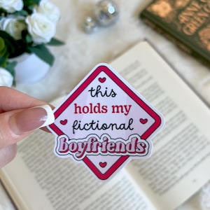 Puede incluir: Una pegatina de forma de diamante rosa y blanca con el texto "this holds my fictional boyfriends" en blanco y negro. La pegatina tiene pequeños corazones rosas alrededor de los bordes.