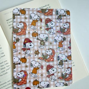 Puede incluir: Una pegatina rectangular con un patrón repetitivo de personajes de Snoopy con ropa de otoño, incluyendo gorros, bufandas y jerséis, sosteniendo calabazas, libros y hojas sobre un fondo de cuadros beige con un efecto de estrellas holográficas.