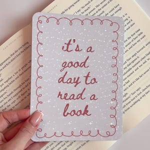 Peut inclure: Une carte rectangulaire avec le texte "It's a good day to read a book" dans un style manuscrit, entourée d'une bordure rouge. Le fond est gris clair avec des paillettes.