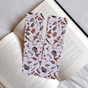 Peut inclure: Deux marque-pages blancs avec un motif répété d'illustrations sur le thème d'Harry Potter. Les illustrations comprennent un train express d'Hogwarts, un vif d'or, un chapeau de sorcier, une baguette magique, une paire de lunettes et une fiole de potion. Le fond est blanc avec un contour rose clair autour de chaque illustration.