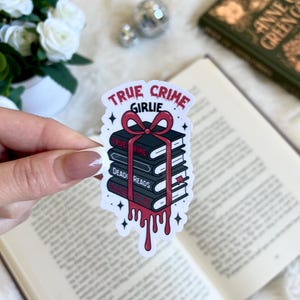 May include: A white sticker with a stack of books tied with a red ribbon. The sticker says "True Crime Girlie" and the books have titles like "True Crime" and "Dead Reads". Red paint drips from the ribbon.