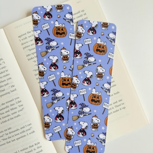 Può includere: Due segnalibri rettangolari in carta con sfondo viola chiaro caratterizzati da un motivo ripetuto di Snoopy in vari costumi di Halloween, tra cui con cappello da strega su una scopa, mantello da vampiro e seduto su zucche arancioni con cartelli BOO.