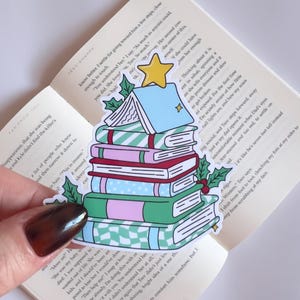 Puede incluir: Una colorida pegatina con forma de árbol de Navidad hecha de libros apilados. Los libros tienen varios patrones y colores, incluyendo verde, rosa y azul. Una estrella amarilla corona el árbol, y ramitas de acebo están a los lados. La pegatina está sobre un libro abierto.