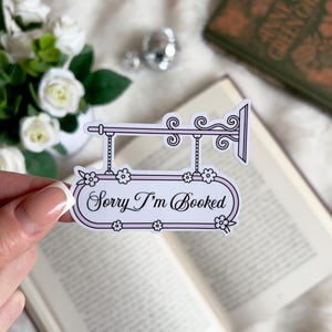 Pode incluir: Um autocolante rosa claro com o texto "Sorry I'm Booked" em itálico. O autocolante tem uma borda branca e detalhes florais. É segurado em frente a um livro aberto e um buquê de flores brancas.