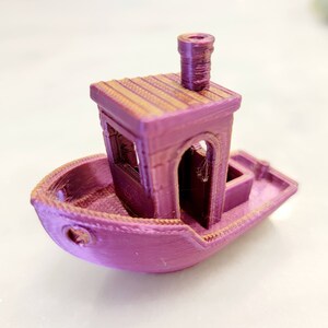 May include: A small, pink, 3D printed toy boat with a cabin and a chimney. The boat has a detailed design with a textured surface.