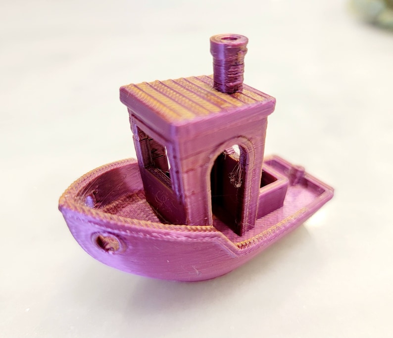 May include: A small, pink, 3D printed toy boat with a cabin and a chimney. The boat has a detailed design with a textured surface.