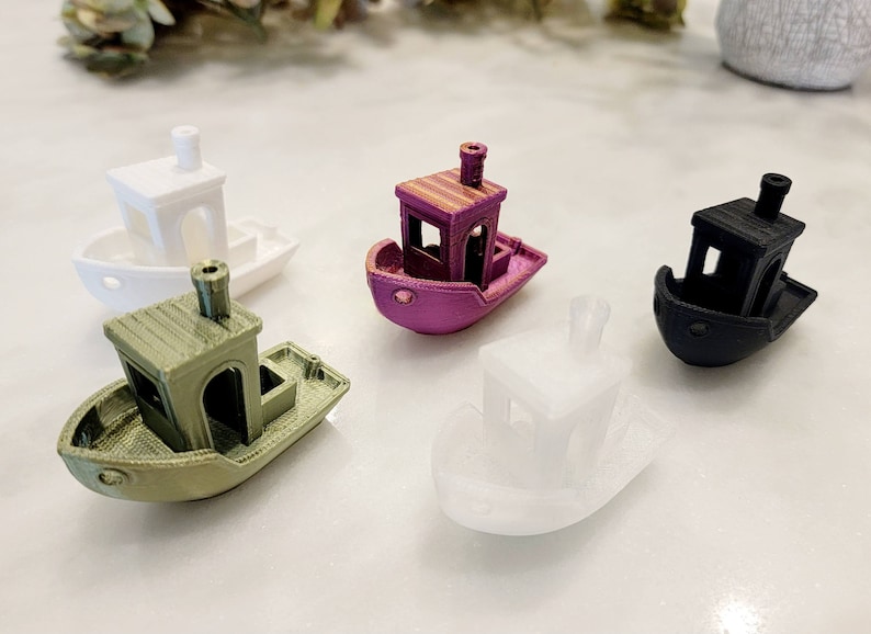 May include: Five miniature wooden boats in different colors, including white, green, purple, black, and white. The boats have a simple design with a small cabin and a flat bottom.