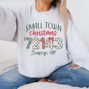 May include: A light gray sweatshirt with the words "SMALL TOWN christmas" in green and red. Below the text are the numbers "72143" with decorative patterns. The text "Searcy, AR" is below the numbers.