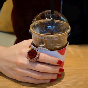 May include: A large, ornate ring featuring a central red gemstone and smaller, colourful surrounding stones. The ring is on a hand with red nail polish, holding a clear plastic cup of iced coffee with a black straw.