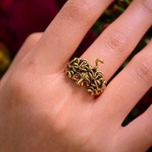 Könnte beinhalten: Ein goldfarbener Ring mit einem detaillierten, strukturierten Design. Der Ring hat einen kleinen, roten Edelstein und einen eingerollten Schwanz. Der Ring wird an einem Finger getragen, mit einem verschwommenen roten und grünen Hintergrund.