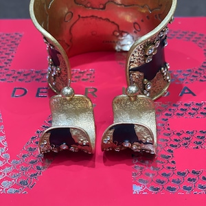May include: A gold-toned cuff bracelet and matching earrings. The cuff features a textured design with black accents and sparkling embellishments. The earrings have a similar design, with a gold top and black base.