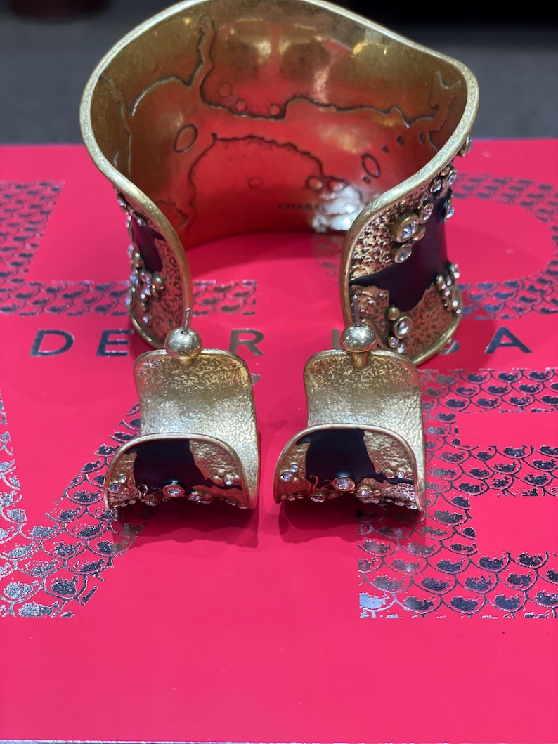 May include: A gold-toned cuff bracelet and matching earrings. The cuff features a textured design with black accents and sparkling embellishments. The earrings have a similar design, with a gold top and black base.