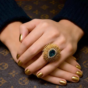 Peut inclure: Une bague dorée avec une grosse pierre centrale de couleur teal et un design en forme d'éclat de soleil. La bague est sertie de petites pierres claires. Les mains, avec du vernis à ongles doré, reposent sur une surface marron à motifs.