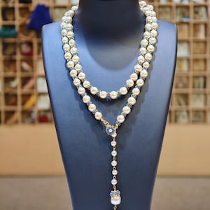 May include: A two-strand pearl necklace with blue accents and a pendant. The necklace features cream-colored pearls and small blue beads. The pendant has a floral design with blue gemstones and a large pearl drop. Displayed on a black mannequin.