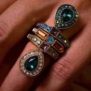May include: A selection of gold rings set with colourful gemstones. The rings display emerald green, sapphire blue, and amethyst purple stones. Some rings have a teardrop shape, while others are bands with smaller gems.