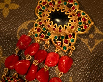 May include: A gold-toned floral brooch with red and green gemstones and a black center. Dangling from the brooch are strands of red coral beads. The brooch has a vintage aesthetic.