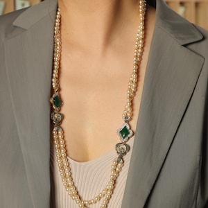 May include: A long, layered pearl necklace with decorative gold and green accents. The necklace features two strands of pearls, with emerald-colored, diamond-shaped and heart-shaped embellishments. The necklace is worn over a neutral-colored top and a gray blazer.