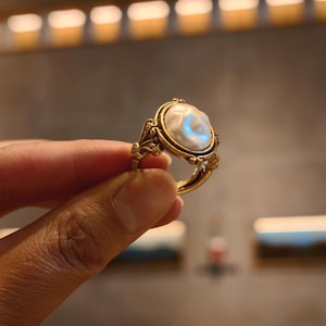May include: A gold-toned ring featuring an oval, off-white pearl. The ring has an ornate, filigree design around the pearl and on the band. The pearl has a subtle, iridescent sheen. The ring is held up against a blurred background.