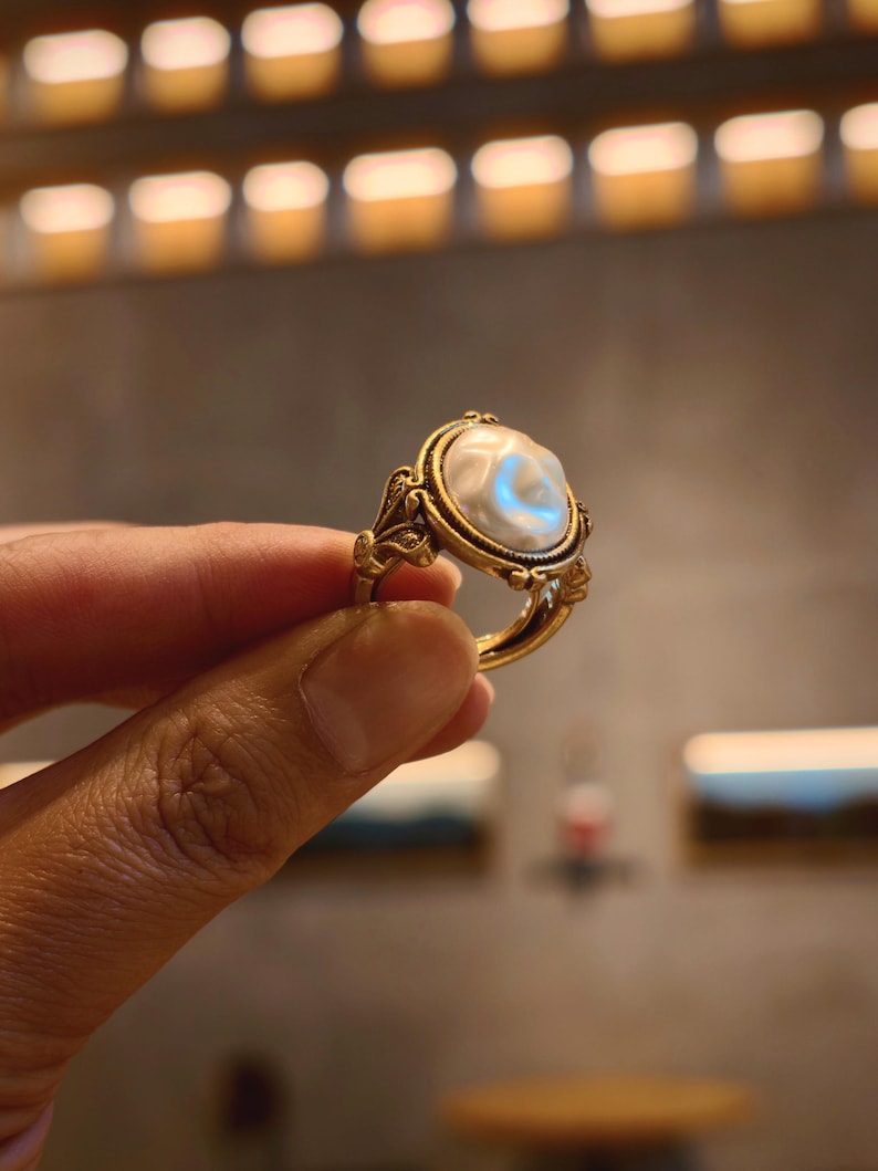 May include: A gold-toned ring featuring an oval, off-white pearl. The ring has an ornate, filigree design around the pearl and on the band. The pearl has a subtle, iridescent sheen. The ring is held up against a blurred background.