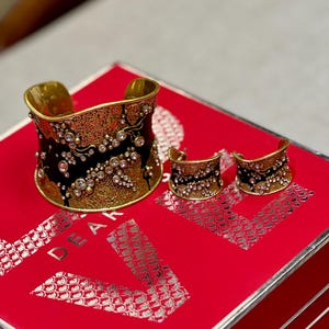 May include: A gold-toned cuff bracelet and matching earrings set. The jewelry features a black enamel design with floral accents and clear crystal embellishments. The set is displayed on a red surface.