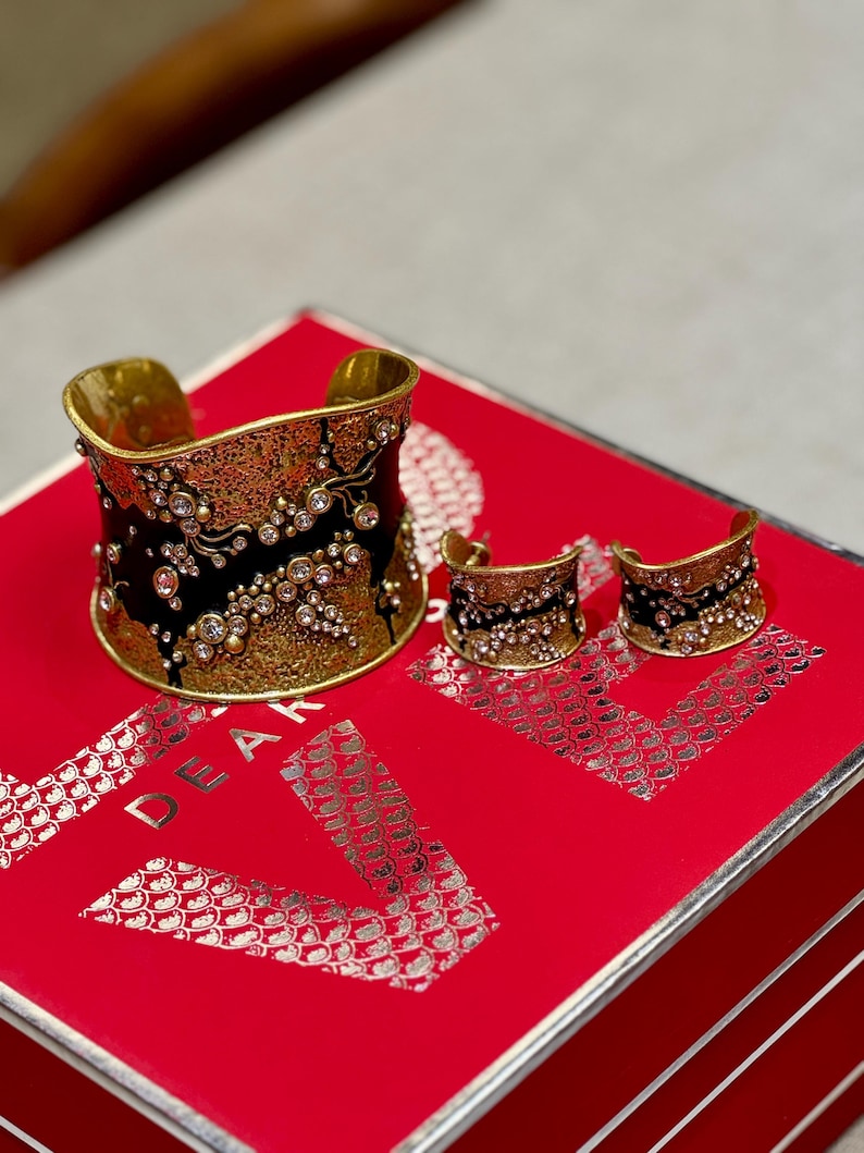 May include: A gold-toned cuff bracelet and matching earrings set. The jewelry features a black enamel design with floral accents and clear crystal embellishments. The set is displayed on a red surface.