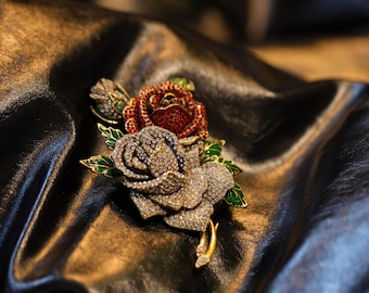 May include: A decorative brooch in the shape of three roses. The largest rose is silver-toned and covered in crystals. The other two roses are red and silver, with green and gold detailing on the leaves and stem. The brooch is set against a dark, textured backdrop.