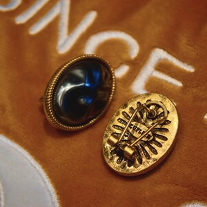 May include: Two vintage oval-shaped brooches. One brooch features a dark, glossy stone set in a gold-toned frame with a rope-like border. The other brooch has a gold-toned back with a pin closure and the word "ZUPLI".