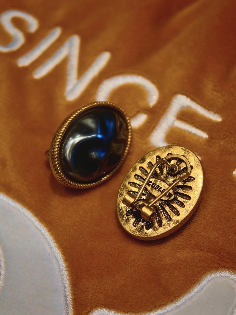 May include: Two vintage oval-shaped brooches. One brooch features a dark, glossy stone set in a gold-toned frame with a rope-like border. The other brooch has a gold-toned back with a pin closure and the word "ZUPLI".