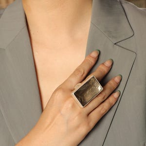 May include: A large, rectangular silver ring. The ring features a thick band and a prominent, rectangular face with a reflective, textured surface. The ring is a statement piece, likely made of metal, and is worn on a finger. The ring's design is modern and geometric. Adjustable comfort fit — each ring contains a discreet inner mechanism that gently adapts to your finger size.