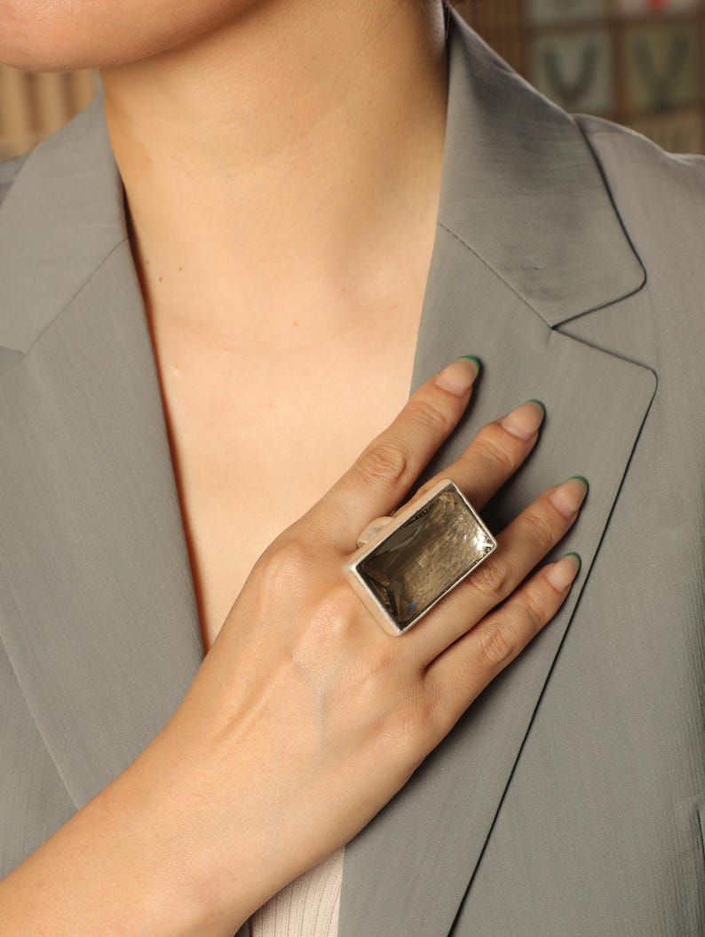 May include: A large, rectangular silver ring. The ring features a thick band and a prominent, rectangular face with a reflective, textured surface. The ring is a statement piece, likely made of metal, and is worn on a finger. The ring's design is modern and geometric. Adjustable comfort fit — each ring contains a discreet inner mechanism that gently adapts to your finger size.