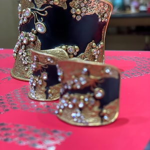 May include: Two ornate, gold-toned metal cuffs with black enamel and sparkling crystal accents. The larger cuff is wider, while the smaller one is a ring. Both have a textured, organic design.