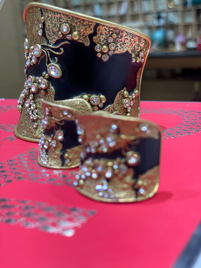 May include: Two ornate, gold-toned metal cuffs with black enamel and sparkling crystal accents. The larger cuff is wider, while the smaller one is a ring. Both have a textured, organic design.