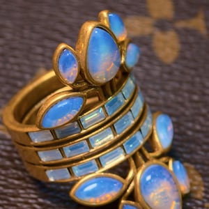 Könnte beinhalten: Ein goldfarbener Ring mit mehreren Bändern, verziert mit hellblauen, ovalen Edelsteinen und rechteckigen, hellblauen Steinen. Der Ring hat ein geschichtetes Design mit floraler Anordnung.