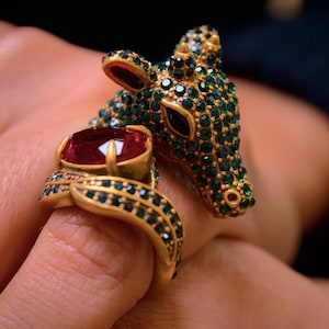 Op de afbeelding: Twee gouden ringen. Eén ring heeft een hertkopontwerp ingelegd met groene edelstenen. De andere ring heeft een grote, ovale, rode edelsteen. Beide ringen hebben gouden banden met groene edelsteenaccenten. De ringen zitten aan een vinger.