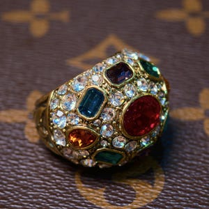 May include: A gold-toned ring encrusted with a variety of colorful gemstones and clear crystals. The ring features a large, oval red stone, a rectangular blue stone, and several other gems in shades of green, orange, and purple. The ring is set against a brown background.