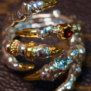 Pode incluir: Um anel prateado com um design em forma de garra. As pontas das garras são douradas, e uma garra apresenta uma pequena gema vermelha e redonda. O anel tem um aspeto metálico texturizado.