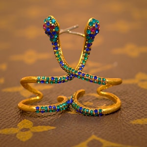 May include: Gold-toned snake-shaped earrings with blue, green, and pink gemstones. The earrings are designed in a stylized snake form, with the gemstones adding color and sparkle. The earrings are set against a brown background.