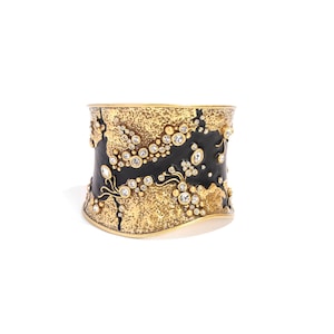 May include: A wide, gold-toned cuff bracelet with a textured, black and gold design. The bracelet is embellished with numerous clear, round gemstones. The words "GLOBAL JEWELRY" are printed on the bottom left.