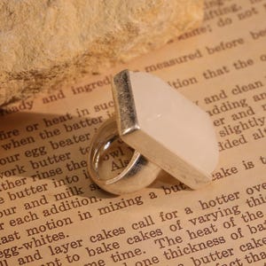 May include: A silver ring with a rectangular, light-colored stone. The ring has a modern design, with a thick band and a flat, geometric stone. The ring is set against a backdrop of a vintage book page.