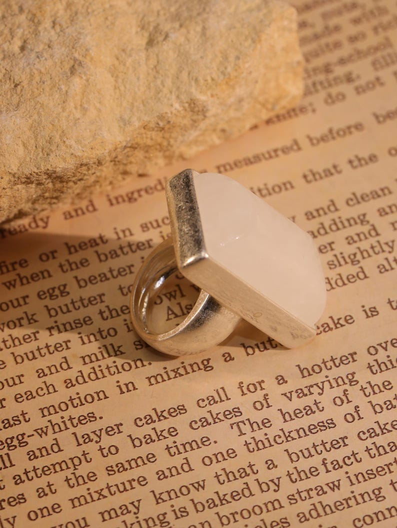 May include: A silver ring with a rectangular, light-colored stone. The ring has a modern design, with a thick band and a flat, geometric stone. The ring is set against a backdrop of a vintage book page.
