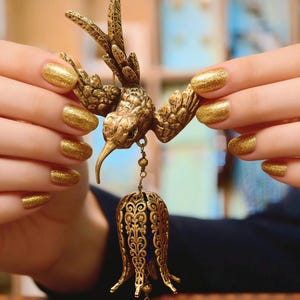 May include: A detailed gold-toned hummingbird brooch with outstretched wings, holding a blue and gold filigree pendant with a blue bead. The brooch is held up by hands with gold glitter nail polish.
