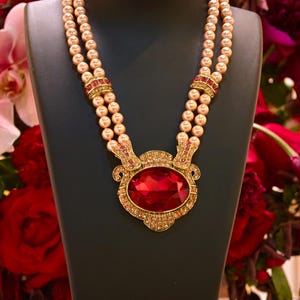 May include: A pearl necklace with a large, oval, red gemstone pendant. The necklace has three strands of pearls and gold-colored accents with red gemstones. The jewelry is displayed on a black mannequin.