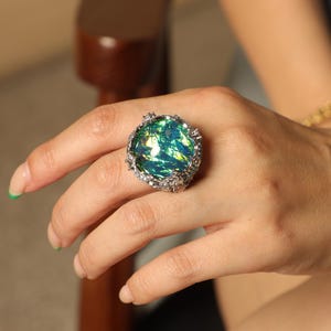 May include: A large, ornate ring featuring a vibrant, iridescent green and blue stone. The stone is set in a silver-toned metal with a detailed, textured design around the edges. The ring is a statement piece, perfect for adding a pop of color.